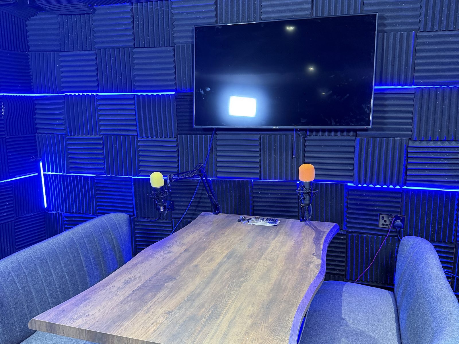 The podcast studio: acoustic foam walls, blue LED strip and two boom microphones at a timber table